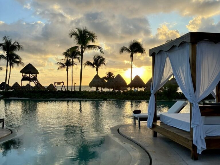 Tropical sunset over a serene pool, with a white-canopied daybed in the foreground, palm trees, and straw huts silhouetted against a dramatic sky.