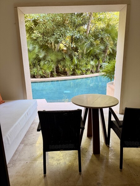 A serene patio overlooking a turquoise pool, bordered by lush greenery. Two wicker chairs face a small round table, with a white sofa on the left. Calm, inviting setting.