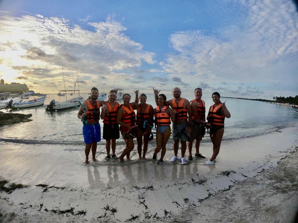 A group of eight people in orange life jackets stands on a beach at sunset. They smile and pose with snorkels, with boats and calm water in the background.