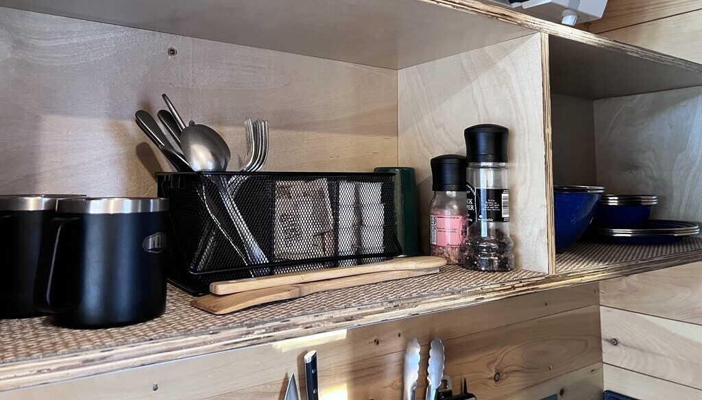 Wooden shelf with black mugs, a metal cutlery holder, spice shakers, and blue bowls. An air conditioning unit is mounted above, conveying a tidy kitchen setup.