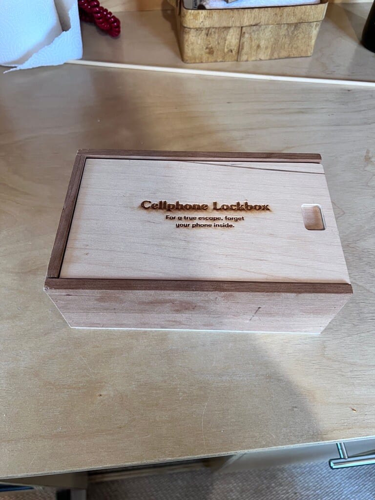 Wooden lockbox labeled "Cellphone Lockbox" sits on a table, suggesting a playful way to disconnect. Warm, rustic tone emphasizes simplicity and escape.