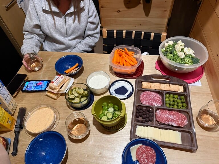 A table spread with a charcuterie board, crackers, salami, cheese, olives, and veggies in bowls. Person holding a glass, Cozy, inviting setting.