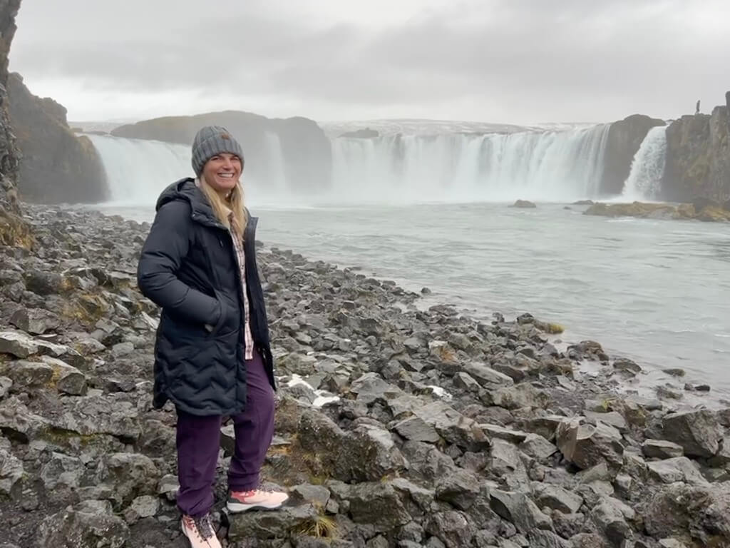 A woman in a black coat and gray beanie stands on a rocky shore, smiling in front of a wide waterfall under a cloudy sky, conveying a sense of adventure.