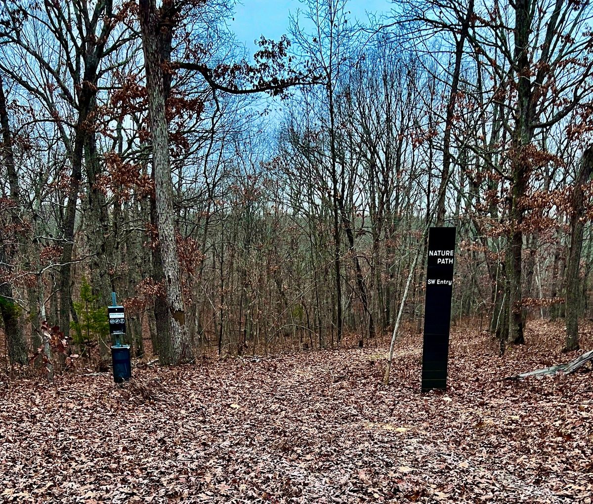 Forest scene with bare trees, scattered leaves covering the ground, and a sign reading "Nature Path SW Entry," suggesting a serene, autumnal setting.