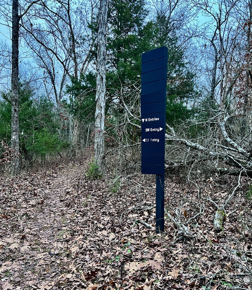 A trailhead in a sparse forest features a dark signpost directing to “N Entries,” “SW Entry,” and “SE Entry.” The ground is covered in dry leaves.