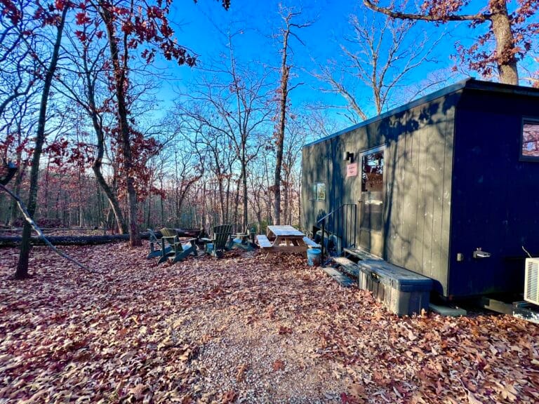 Cabin in a forest surrounded by autumn leaves. Adirondack chairs and a picnic table are nearby, under a clear blue sky. Peaceful and rustic setting.
