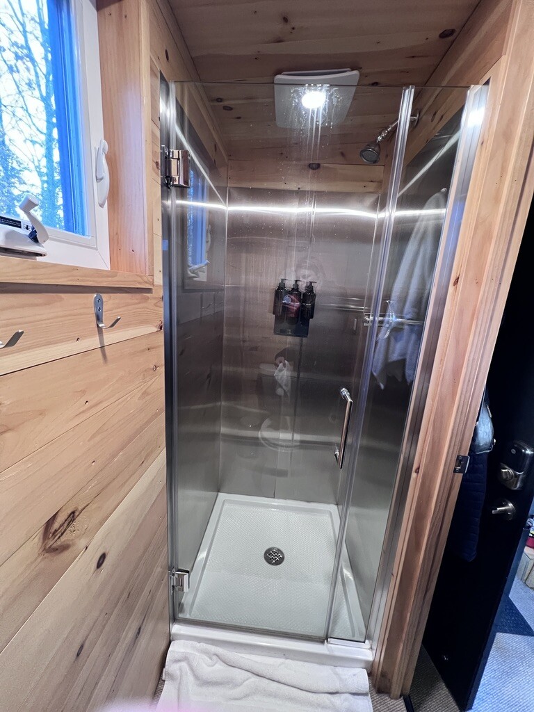 A small glass shower with metal fixtures is set in a wooden cabin-like bathroom. A towel hangs on the wall, and a window lets in natural light.