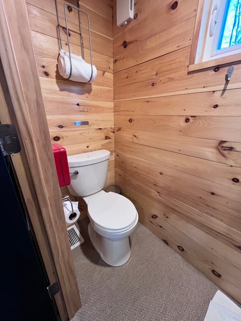 Small rustic bathroom with wooden walls featuring a white toilet. Towels hang above in a metal rack. The space feels cozy and minimalist.