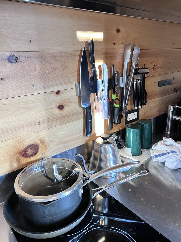 Kitchen countertop with a wooden backsplash. Knives, tongs, and kitchen tools are mounted on a magnetic strip. Pots, pans, and a kettle rest on the counter, creating an organized and functional space.