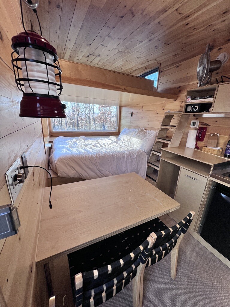 Cozy cabin interior with wooden walls and ceiling. A bed with white linens sits by a large window showing trees. Nearby, a kitchenette and wooden table.