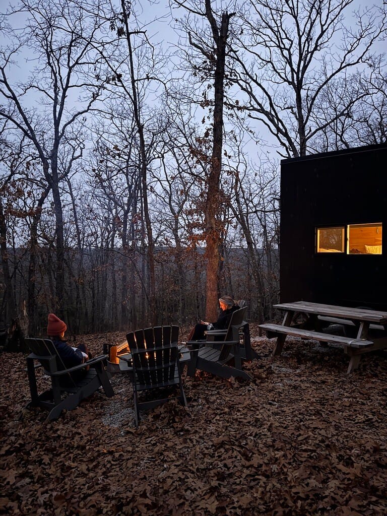 Two people in winter hats sit by a lit fire pit among leaf-covered ground and bare trees, next to a dark cabin at twilight, conveying tranquility.