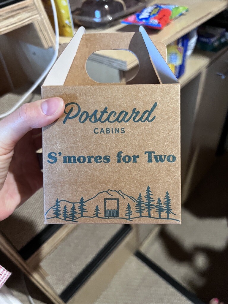 A hand holds a brown takeout box labeled "Postcard Cabins S'mores for Two" with a mountain and trees illustration, evoking a cozy, outdoor vibe.