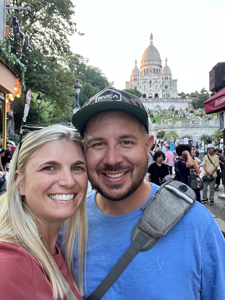 A smiling couple takes a selfie in front of the Sacré-Cœur Basilica in Paris. The scene is vibrant with tourists and greenery, evoking joy and travel excitement.