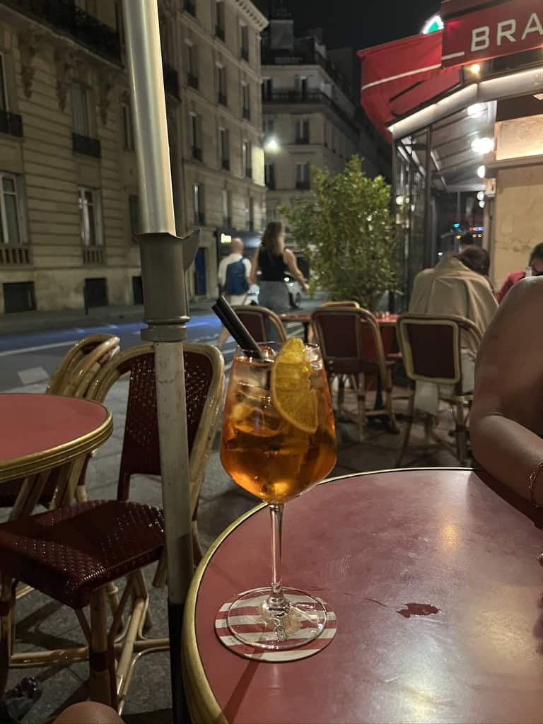 A refreshing Aperol Spritz with lemon slice on a red outdoor table in a lively Paris street at night.