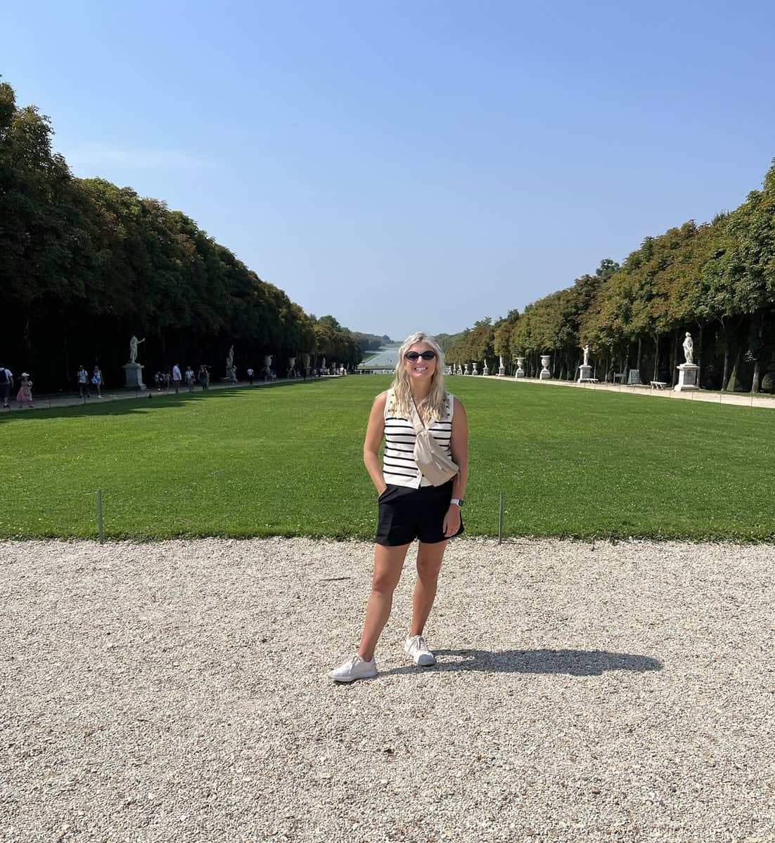Woman standing in the gardens of Versailles with trees and statues in the background.
