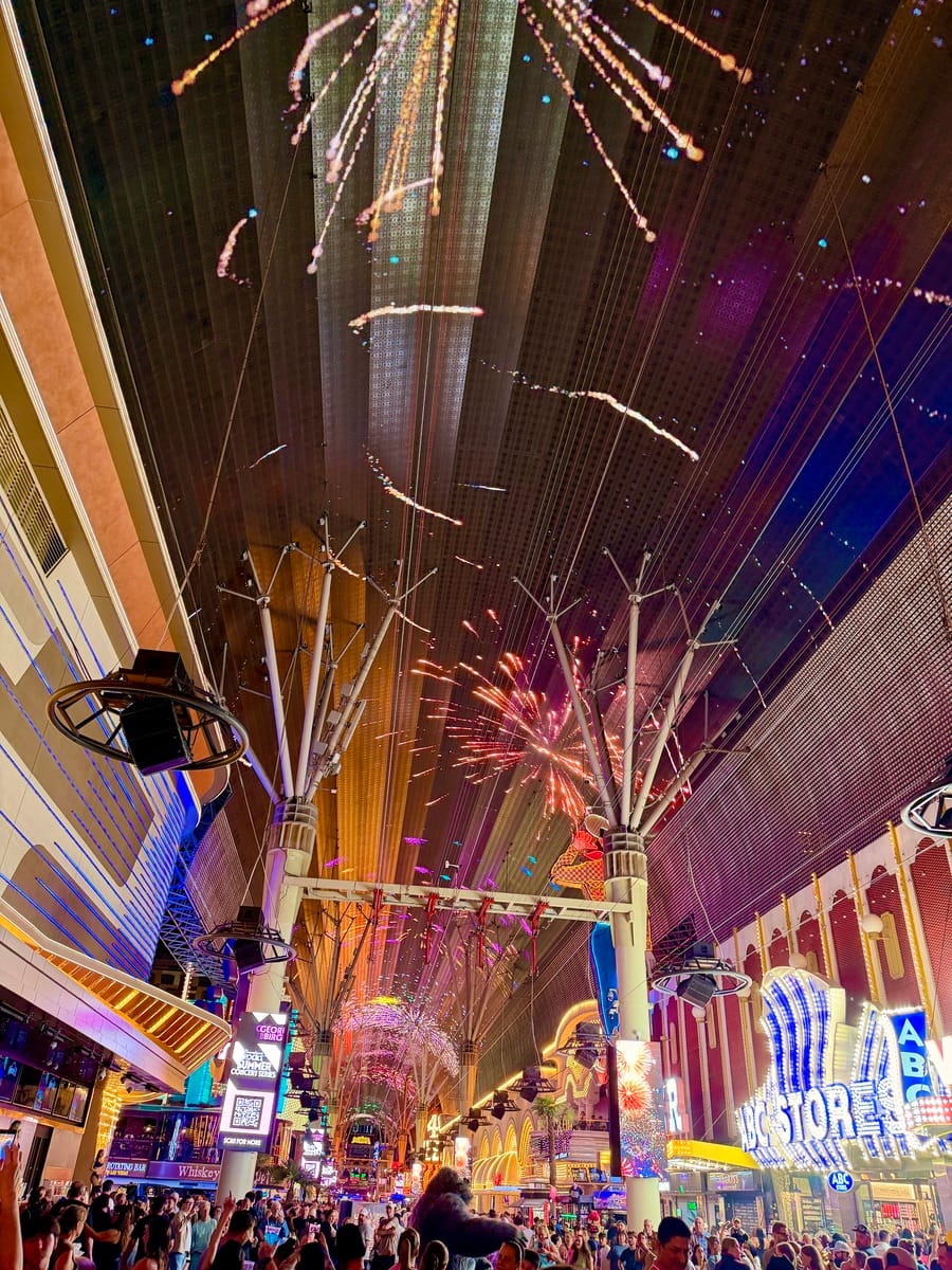 Fireworks display over a lively city street at night, with bright lights and colorful explosions.