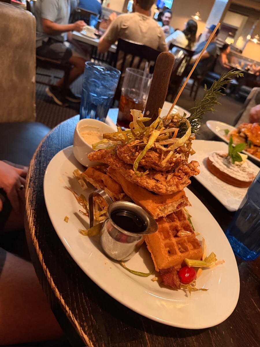 Crispy fried chicken and waffles with syrup and garnishes at a lively restaurant.
