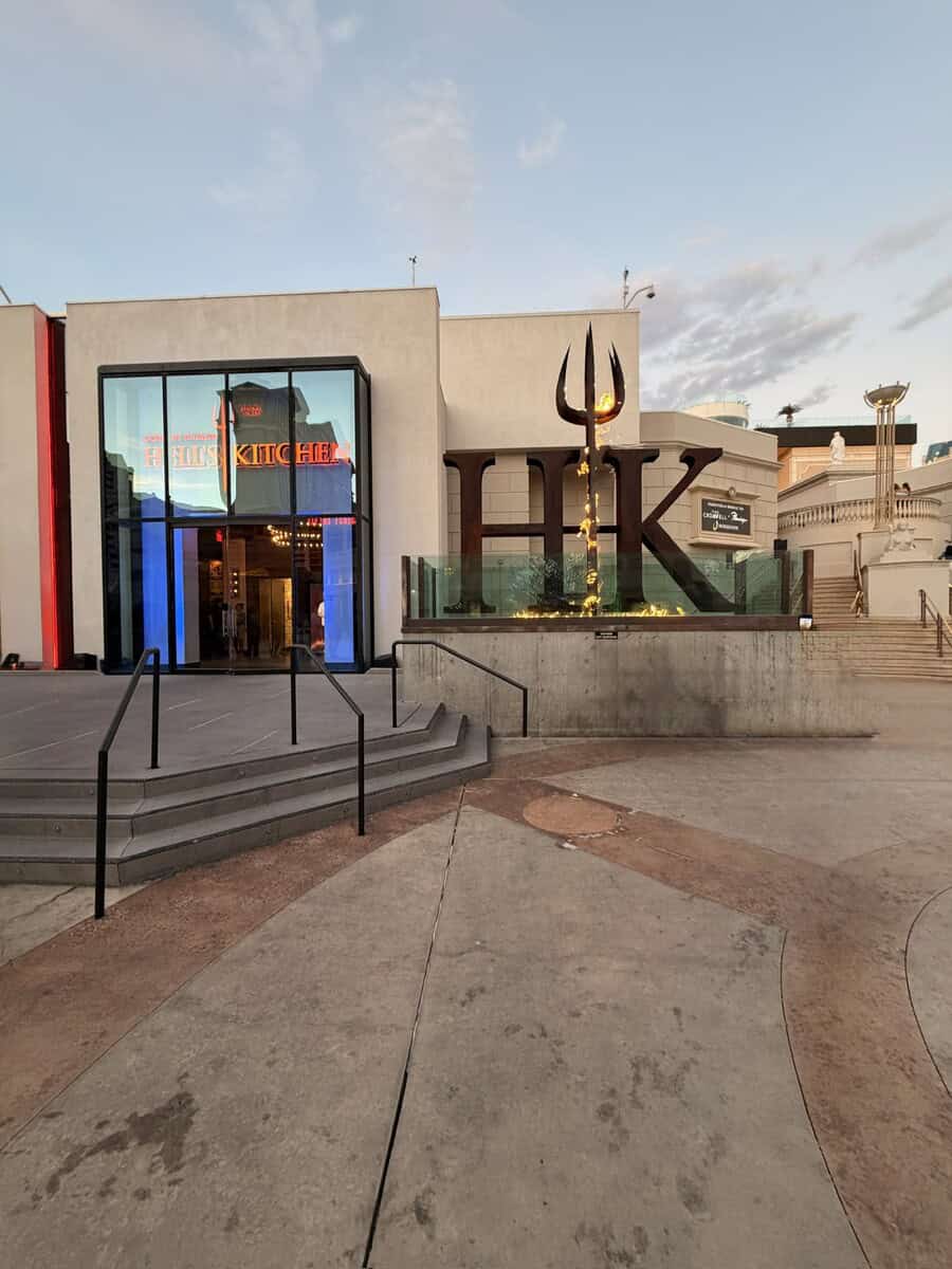 Brightly lit exterior of a modern shopping mall with a large "K" sculpture and a trident symbol, sho.