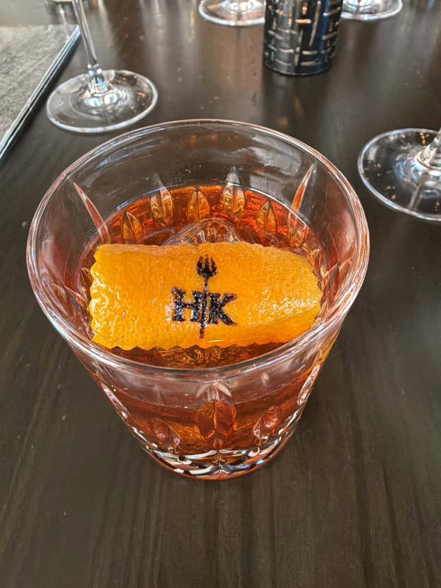 A colorful cocktail with an orange garnish featuring the HK logo, served in a glass on a dark table.
