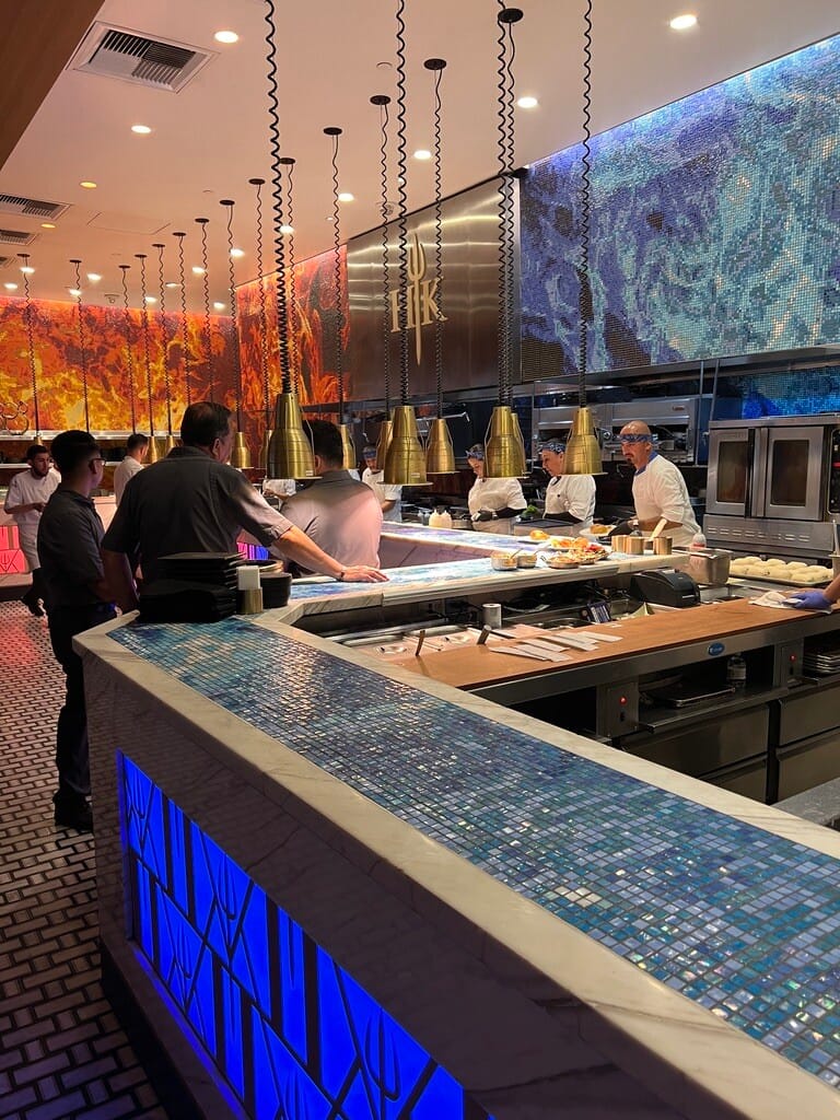 Open kitchen with chefs preparing dishes under warm pendant lights. Vibrant mosaic walls depict fire and water, creating a dynamic atmosphere.