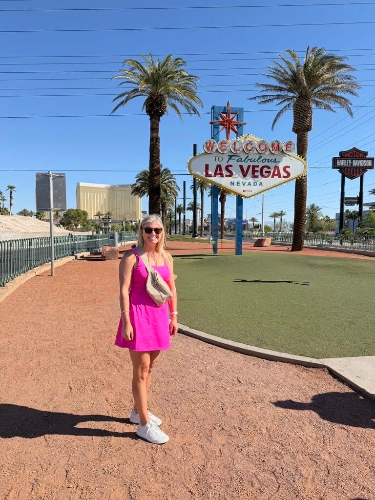 Bright woman in pink dress standing near Las Vegas Welcome sign on sunny day.