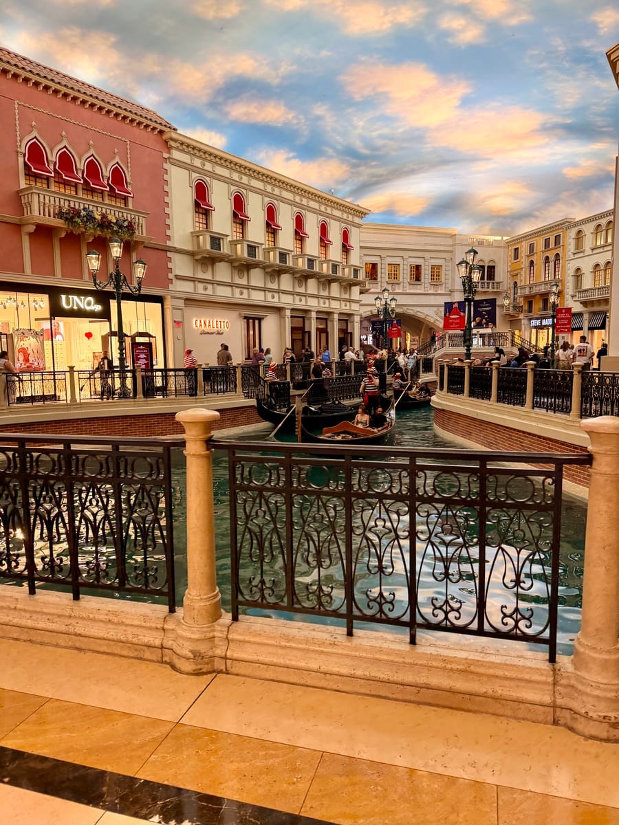 Indoor Venetian-themed shopping mall with a canal, gondolas, and ornate buildings under a painted sky. Shoppers and gondoliers add lively ambiance.
