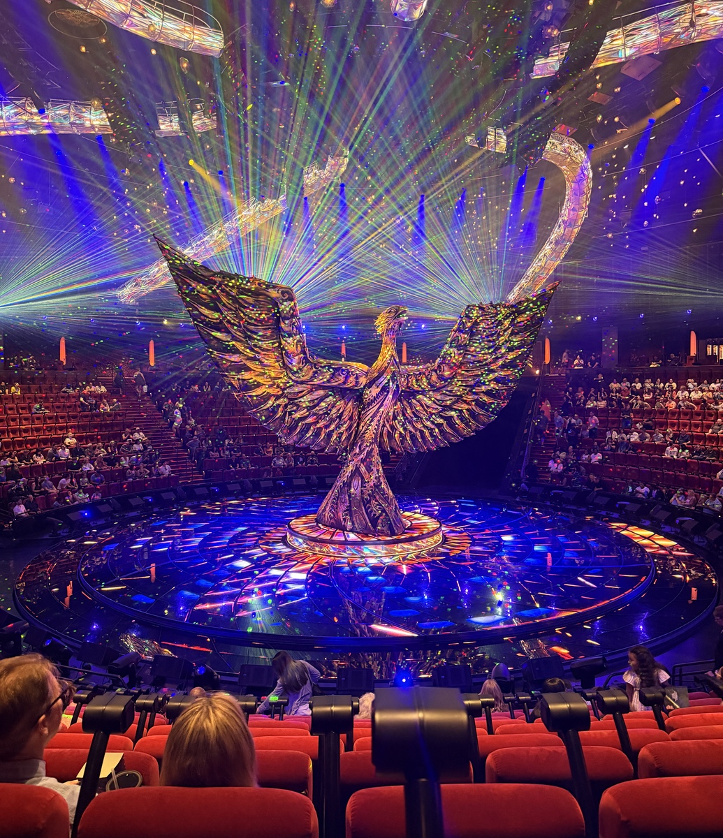 A vibrant, illuminated phoenix sculpture dominates the stage in a theater, surrounded by colorful laser lights and an audience seated in red chairs.