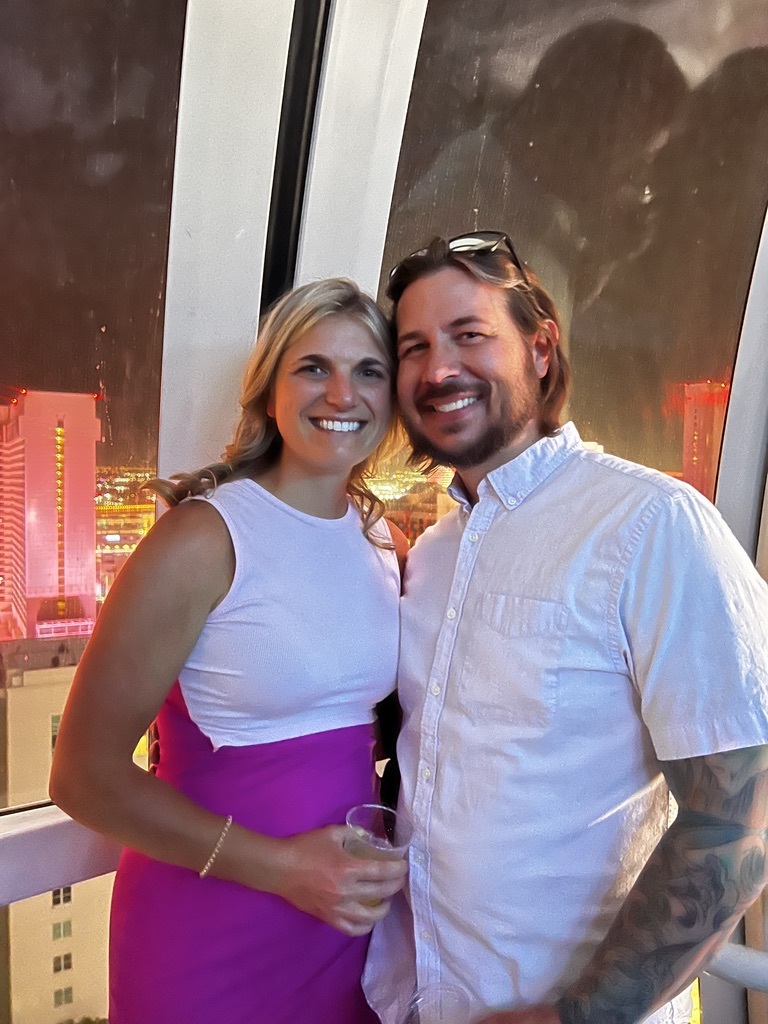 Smiling woman in a white and pink dress stands with a man in a white shirt at night, inside a glass-walled enclosure with city lights behind them.