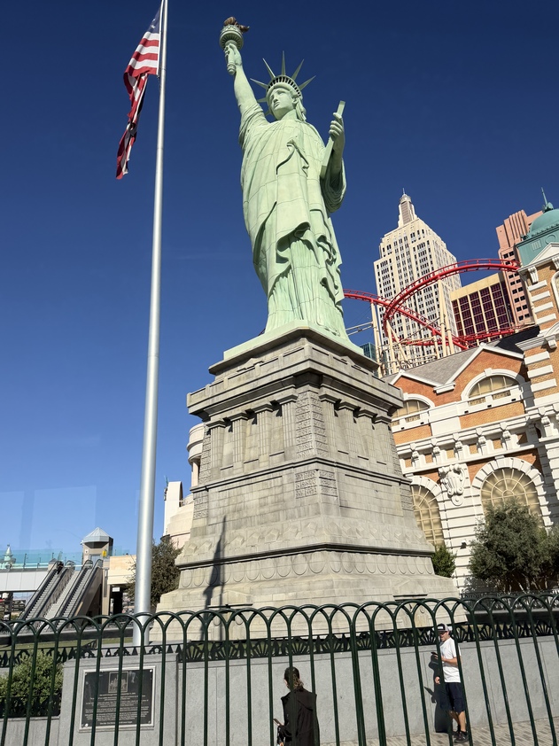 Replica of the Statue of Liberty in bright sunlight, surrounded by buildings and a roller coaster. A flag flutters nearby, creating a vibrant urban scene.