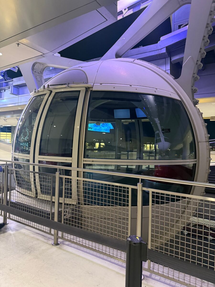 Large glass-enclosed Ferris wheel cabin at a station, illuminated by soft blue and purple lights. A metal fence surrounds the area.