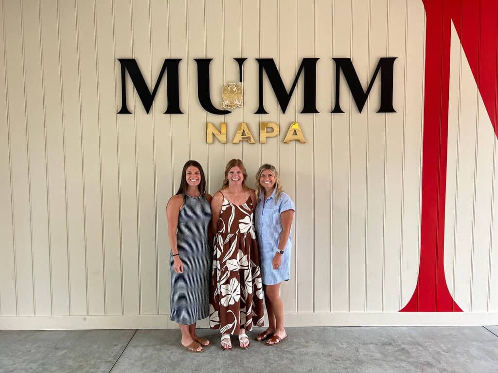 Three women standing in front of Mumm Napa sign at winery.