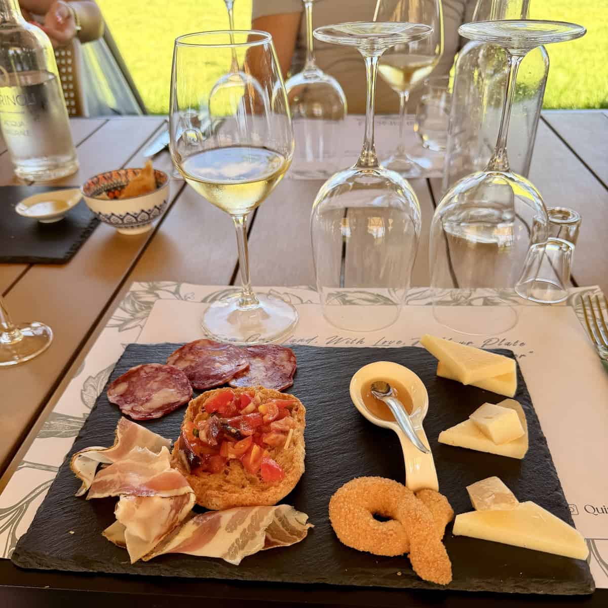 Cheese, cured meats, and bread on a slate platter with wine glasses in the background.