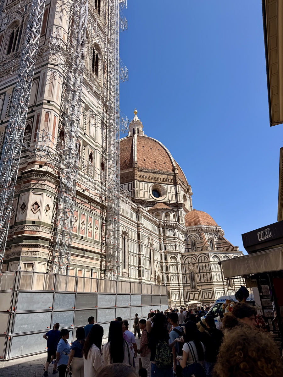 Florence’s Cathedral of Santa Maria del Fiore rises above a busy street, its ornate marble façade, red-tiled dome, and crowds set against a clear blue sky.