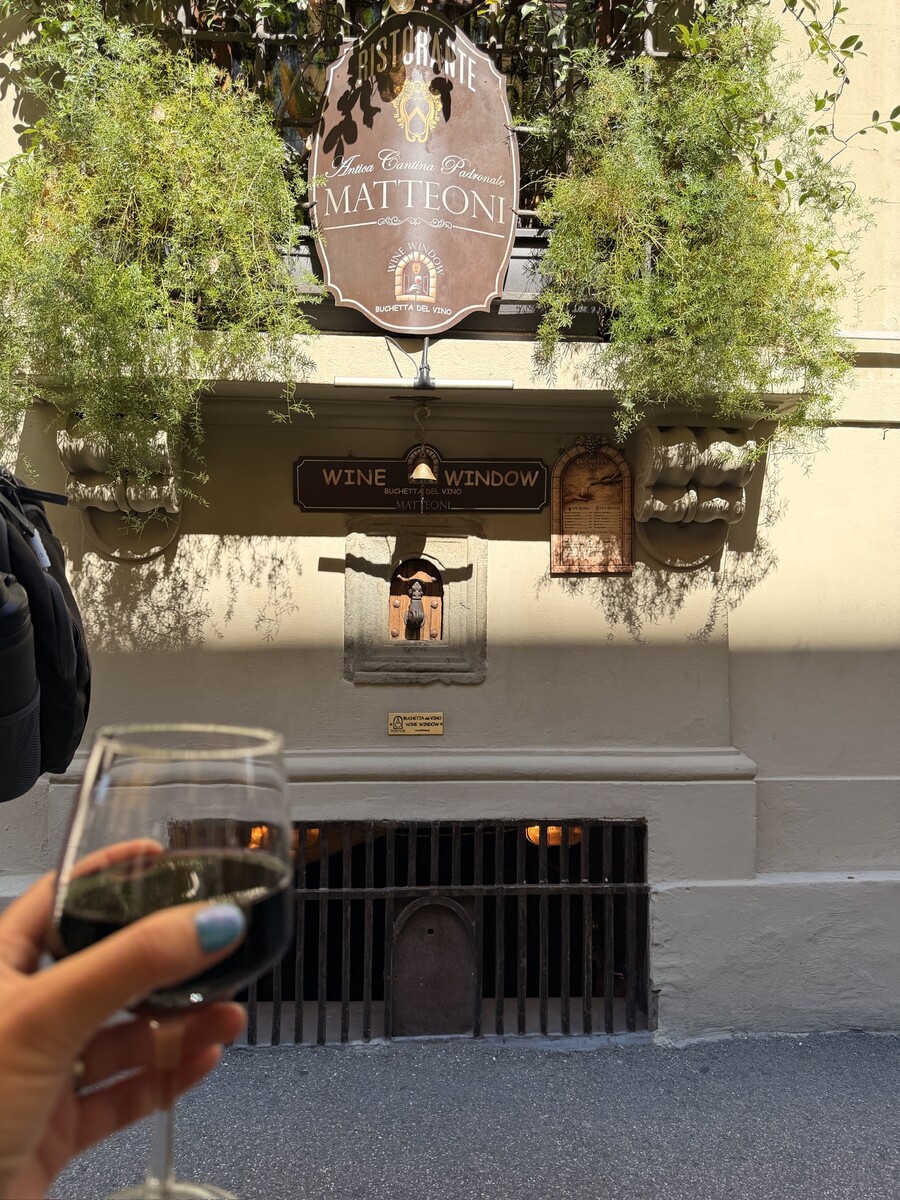 A hand holds a glass of red wine outside a sunlit Napa Valley tasting room, framed by leafy vines and signs for wine tasting; warm, relaxed atmosphere.