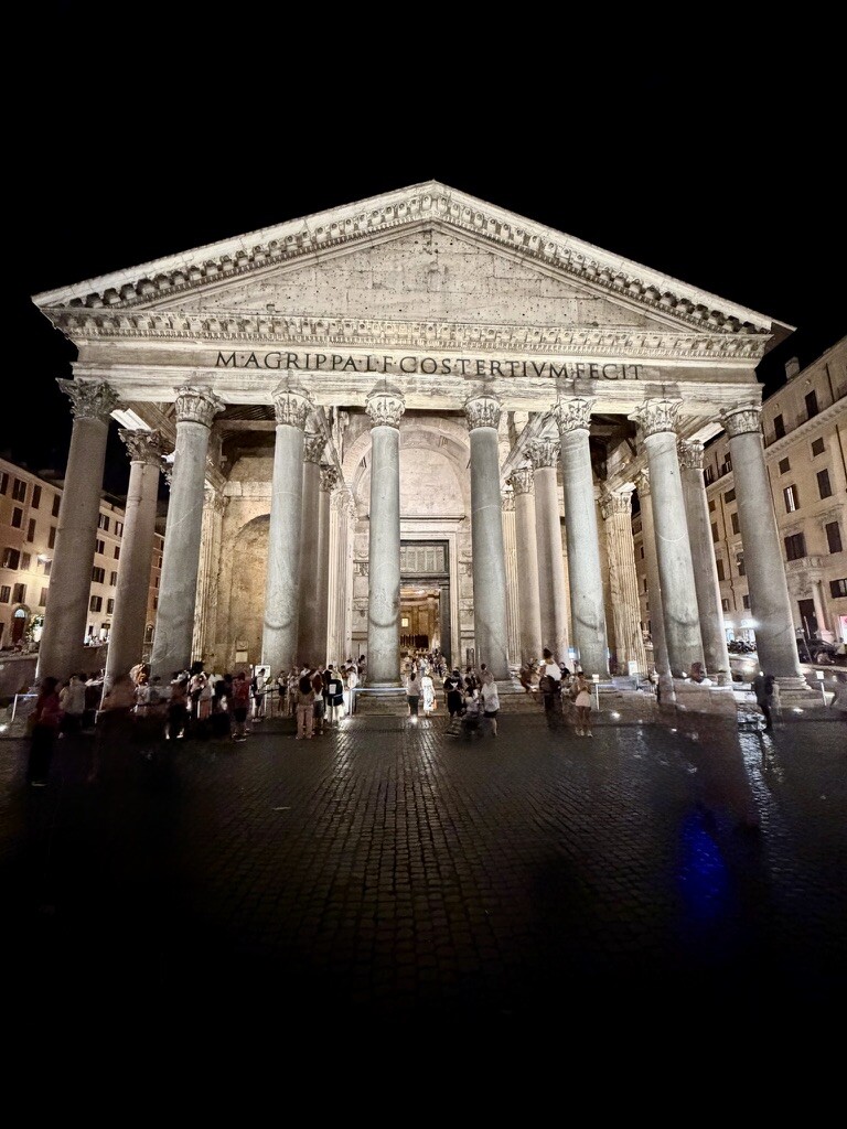 Night view of Rome’s Pantheon, its illuminated ancient columns and triangular pediment towering over visitors gathered in the wet piazza.
