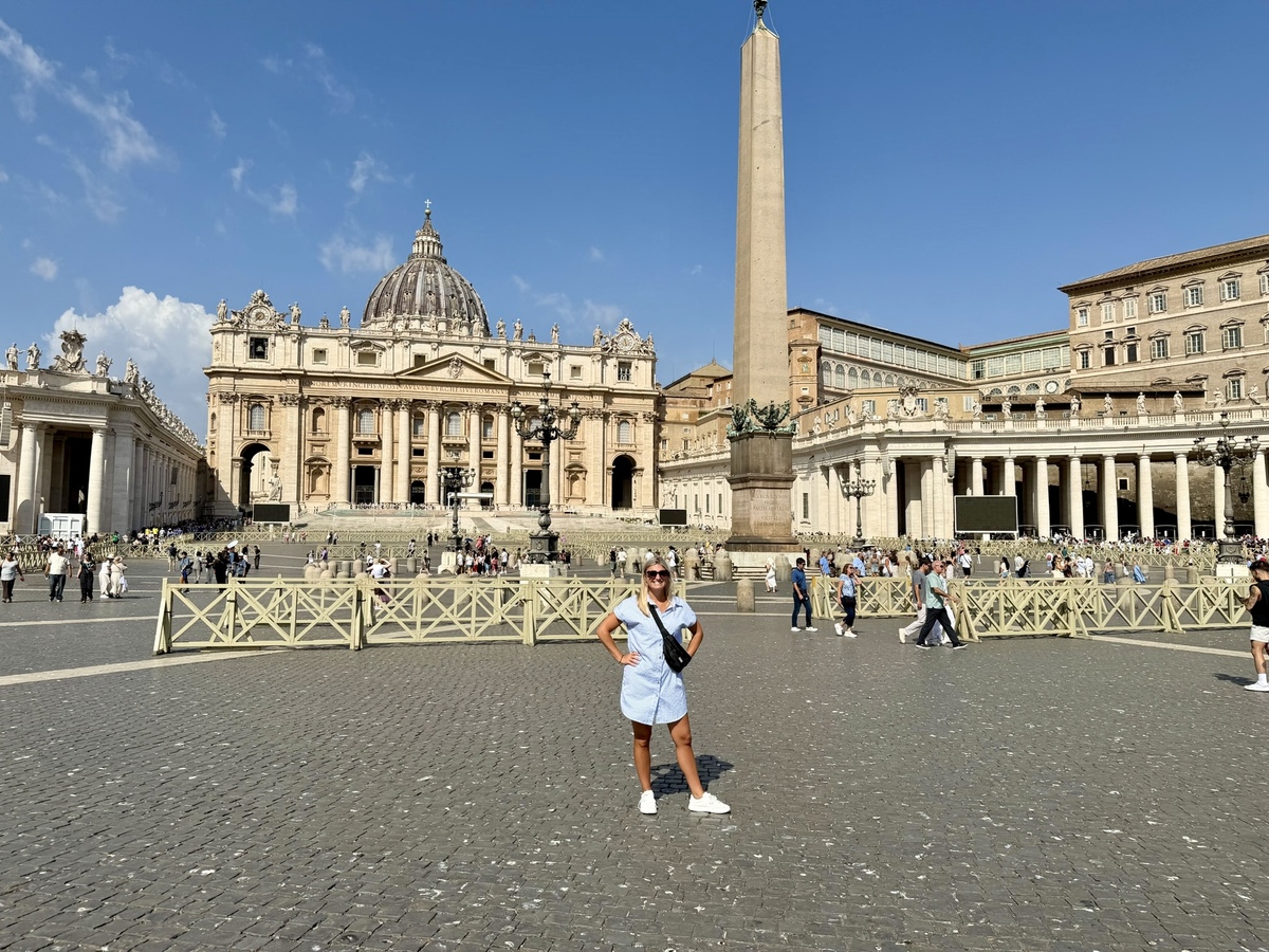 Brenda poses in St. Peter’s Square before St. Peter’s Basilica and its central obelisk, with crowds, colonnades, and a bright blue sky.